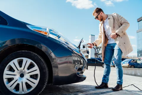 Junger Mann mit Sonnenbrille steckt das Ladekabel in sein Elektroauto.